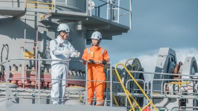 Two maritime workers on ship deck wearing safety gear; one in white coveralls and one in orange.